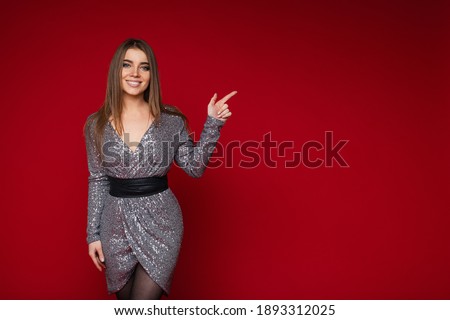 Smiling young female in festive silver dress pointing on blank space for advertising content on red background