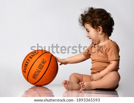 Games for children. Playful curly little boy dressed in a bodysuit sitting barefoot and playing a new basketball on a gray background. Place for text. Concept of active childhood.
