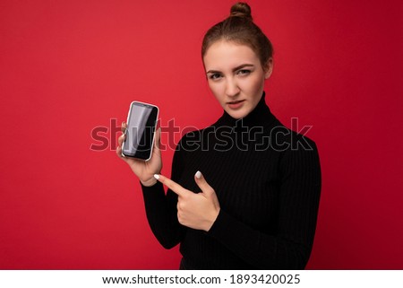 Photo of attractive serious young brunette woman wearing black sweater standing isolated over red background showing mobile phone with empty screen for mockup looking at camera pointing finger at