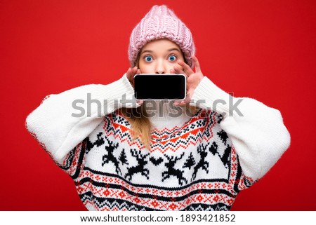 Photo of attractive surprised young blonde woman wearing warm knitted hat and winter warm sweater standing isolated over red background showing smartphone with empty screen for mock up looking at