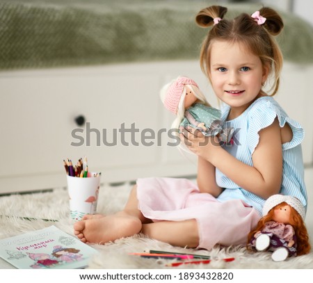 Pretty little kid girl in home dress sit barefooted on soft carpet rug, playing with dolls, drawing with pencils at home. Translation from Russian: Coloring book with stickers puppet world