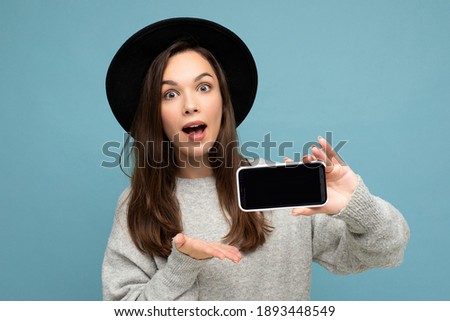 Portrait photo shot of beautiful amazing young woman wearing black hat and grey sweater holding phone showing smartphone isolated on background