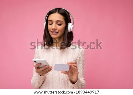 Photo of beautiful happy young brunette woman wearing pink casual sweater isolated over pink background wall wearing white bluetooth wireless headphones and listening to music and using mobile phone