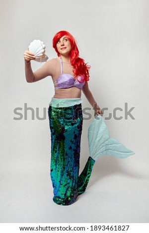 A woman in a mermaid costume with red hair holds a shell in one hand and a tail in the other. White background.
