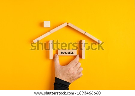 Male hand placing a buy or sell sign in a house made of wooden blocks on yellow background. Buying or selling a hose decision in the real estate market. 