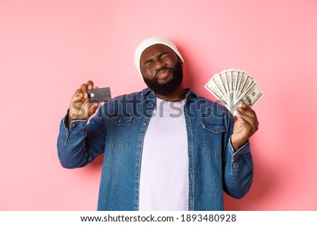 Shopping concept. Sad and whining Black guy showing credit card and dollars, grimacing reluctant, standing over pink background