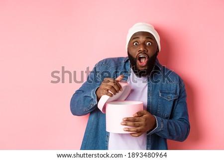 Excited Black man open box with gift and staring at camera grateful and amazed, receiving present, standing over pink background