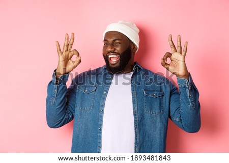 Satisfied african-american man in hipster beanie, showing okay signs in approval, praising good offer and saying yes, smiling pleased, standing over pink background