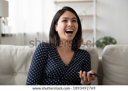 Emotional happy millennial asian mixed race woman holding cell phone in hands, laughing at funny photo video content on social network, enjoying spending relaxed carefree weekend time online at home.