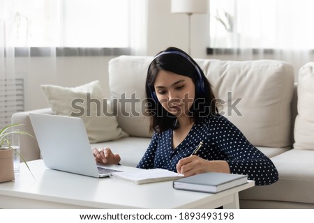 Focused young vietnamese korean woman listening educational lecture on computer using headphones, writing notes in copybook, improving professional knowledge or attending foreign language online class Royalty-Free Stock Photo #1893493198