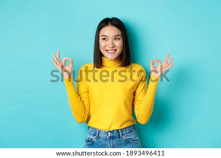 Beautiful japanese girl in yellow sweater, showing Ok signs and smiling, looking left and logo and recommending company, standing over blue background