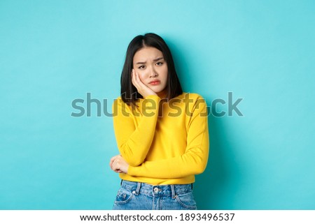 Sad and bored asian girl looking reluctant and unamused at camera, leaning face on hand, standing over blue background