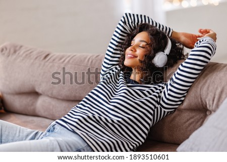 Happy ethnic woman with closed eyes smiling and listening to favorite music in headphones while relaxing on comfortable couch at home Royalty-Free Stock Photo #1893510601
