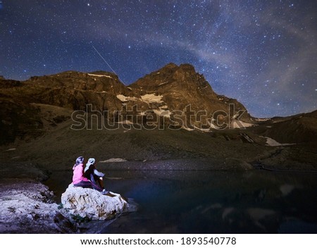 Scenery of mountains with snow at night in Alps. Gorgeous mountain ridge with high rocky peaks, couple sitting on stone near lake, enjoying milky way with shining stars in sky, Matterhorn.