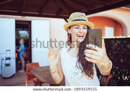 happy trendy mother and child travellers in hats with wheel bags answering incoming video call on a smartphone in the terrace of guest house hotel.