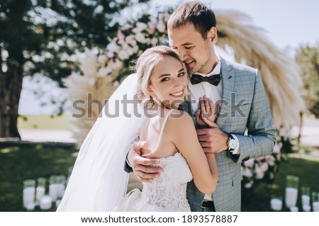 Wedding portrait of smiling newlyweds. Stylish groom in a gray checkered suit and a cute blonde bride in a white lace dress are tenderly hugging in the forest against the background of the arch. Royalty-Free Stock Photo #1893578887