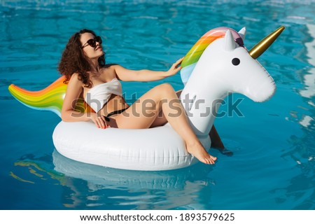 Young beautiful brown-haired curly girl in hat with a good figure tan in the pool on an inflatable unicorn in a white swimsuit