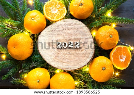 New year's holiday background on a round cut of a tree surrounded by tangerines, live fir branches and golden lights garlands, with wooden numbers date 2022. Citrus aroma, Christmas. Space for text.