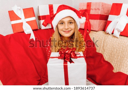 Happy little girl in Christmas sweater and Santa Claus hat, with gifts on the sofa at home, on Christmas morning