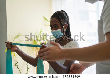 Young African woman wearing a mask doing rehabilitation exercises with a rubber band next to a physiotherapist wearing a mask and a uniform in a clinic Royalty-Free Stock Photo #1893618091