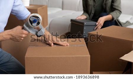 Crop close up of happy couple pack wrap boxes with adhesive scotch moving to new home together. Man and woman renters seal packages with tape dispenser on relocation day. Rent, delivery concept. Royalty-Free Stock Photo #1893636898