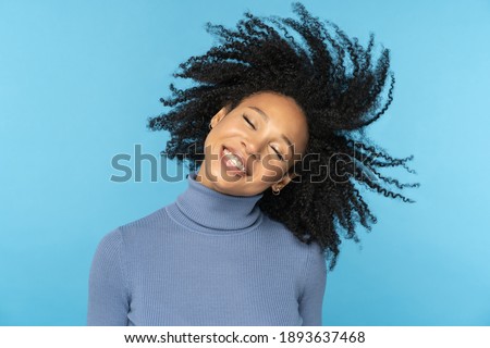 Happy Afro woman smiling, has good mood, dancing with her black curly hair flying, isolated on studio blue background. Excited african american girl with flying hairstyle. Positive life concept.  Royalty-Free Stock Photo #1893637468