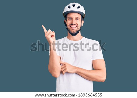 Young handsome man wearing bike helmet with a big smile on face, pointing with hand and finger to the side looking at the camera. 