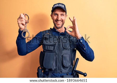 Handsome policeman wearing uniform and bulletprof holding handcuffs over yellow background doing ok sign with fingers, smiling friendly gesturing excellent symbol