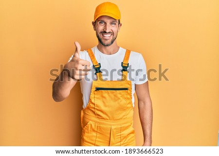 Young handsome man wearing handyman uniform over yellow background doing happy thumbs up gesture with hand. approving expression looking at the camera showing success. 