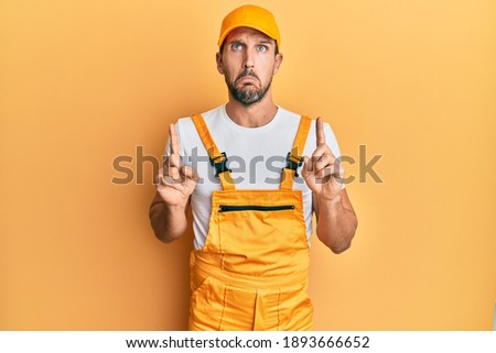 Young handsome man wearing handyman uniform over yellow background pointing up looking sad and upset, indicating direction with fingers, unhappy and depressed. 