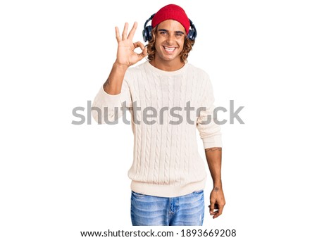 Young hispanic man listening to music using headphones smiling positive doing ok sign with hand and fingers. successful expression. 