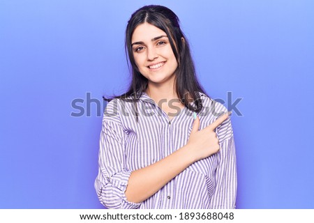 Young beautiful girl wearing casual striped shirt smiling cheerful pointing with hand and finger up to the side 