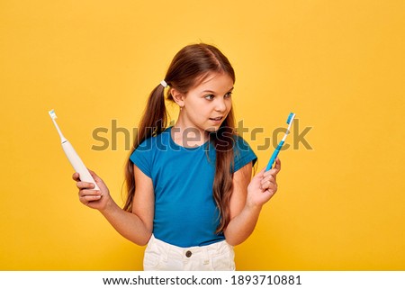 Pretty little girl holds manual and electric sonic toothbrush. Child selects of kind of toothbrush, on yellow background