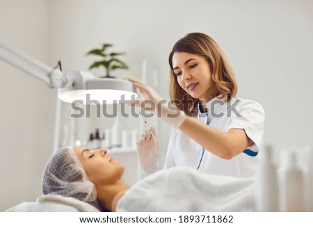 Professional female beautician makes a rejuvenating injection to a young woman who lies relaxed with her eyes closed in a modern beauty salon. Concept of smoothing wrinkles with injections. Royalty-Free Stock Photo #1893711862
