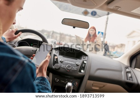 Distracted young male driver looking at the screen of his mobile phone while running over a pedestrian. Technology and transportation concept. Selective focus on hand with smartphone. Horizontal shot Royalty-Free Stock Photo #1893713599