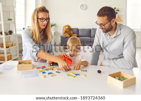 Focused mom, dad and toddler son play and compose words from scattered colored letters. Family sits at home in the living room and spends their free time usefully. Concept of education and development