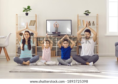 Happy calm young parents teaching their kids to practise meditation. Family with little children sitting cross-legged on warm floor rug at home and learning to do yoga exercise with TV video lesson Royalty-Free Stock Photo #1893725398