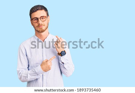 Handsome young man with bear wearing elegant business shirt and glasses in hurry pointing to watch time, impatience, looking at the camera with relaxed expression  Royalty-Free Stock Photo #1893735460