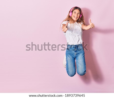 Young beautiful blonde woman smiling happy. Jumping with smile on face listening to music using pink headphones doing ok sign with thumbs up over isolated background