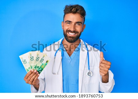 Young hispanic man wearing doctor uniform holding israel shekels smiling happy and positive, thumb up doing excellent and approval sign 