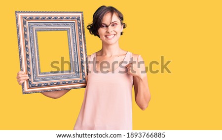 Beautiful young woman with short hair holding empty frame pointing finger to one self smiling happy and proud 