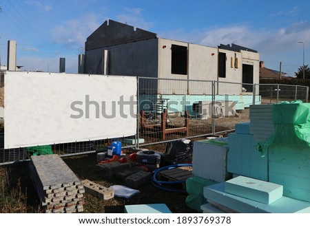 Blank information board on the border of the construction site with a new prefab  house among insulating materials and building equipment. Background for copy space