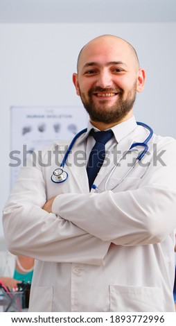Portrait of young man doctor standing in front of camera smiling in medical conference meeting office. Team of specialist doctors working in background talking about symptoms of disease in clinic room