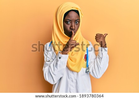 Beautiful african young woman wearing doctor uniform and hijab asking to be quiet with finger on lips pointing with hand to the side. silence and secret concept. 