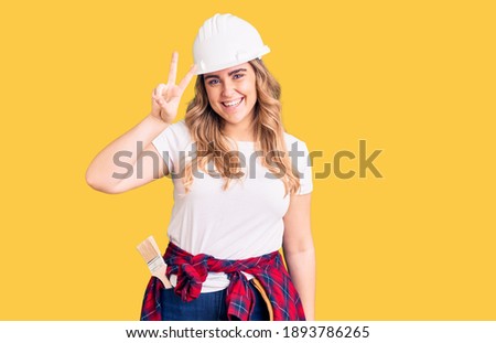Young caucasian woman wearing security helmet showing and pointing up with fingers number two while smiling confident and happy. 