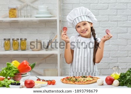 little girl in chef hat and an apron cooking pizza in the kitchen. the child holding sausage slices. having fun