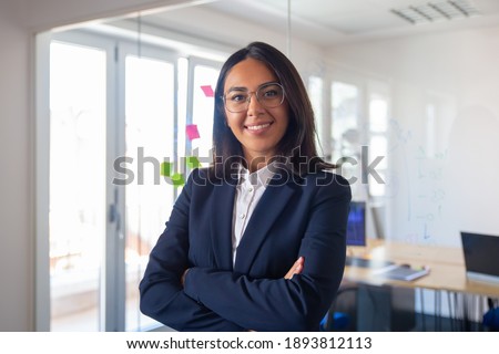 Confident Latin business leader portrait. Young businesswoman in suit and glasses posing with arms folded, looking at camera and smiling. Female leadership concept Royalty-Free Stock Photo #1893812113