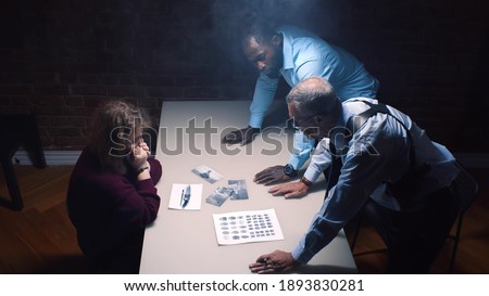 Two police officers and woman criminal in interrogation room. Multiethnic detective showing evidence to female suspect in handcuffs accusing her of crime Royalty-Free Stock Photo #1893830281