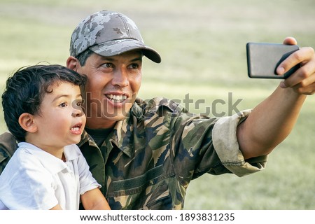 Cheerful disabled military dad and his little son taking selfie together in park. Boy sitting on dads lap. Veteran of war or disability concept