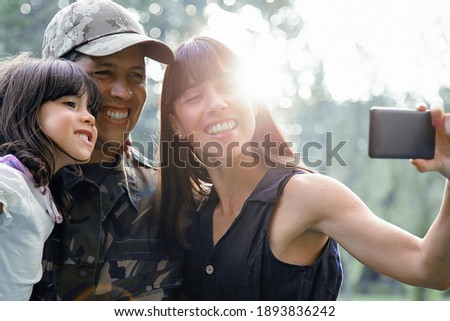 Cheerful military man with wife and little daughter taking selfie on cellphone in city park. Medium shot, sunshine. Family reunion or returning home concept